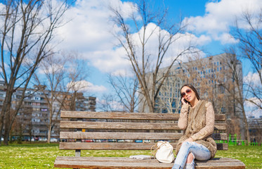 Naklejka na ściany i meble Beautiful young woman sitting at park on bench talking on the smart phone. Blue sky in background. Shallow depth of field.