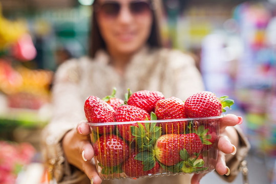 Beautiful young woman holding fresh strawberries at marketplace, focus is on the strawberries. Shallow depth of field.