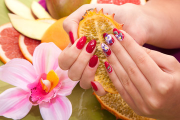 female hand with original red design nails hold a tropical fruit kiwano. Pineapple, grapefruit, mango on the background. Hand care Spa