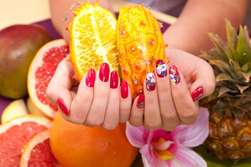 female hand with original red design nails hold a tropical fruit kiwano. Pineapple, grapefruit, mango on the background. Hand care Spa