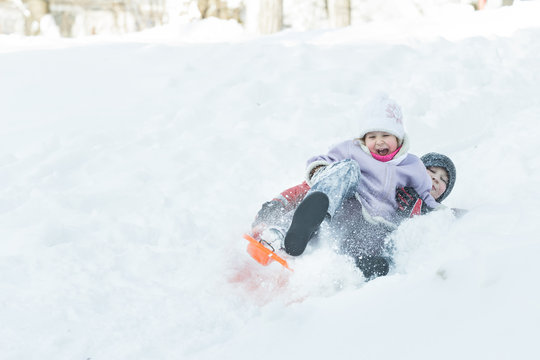 Two children sliding down snowy hill outdoors on orange plastic modern toboggan for kids