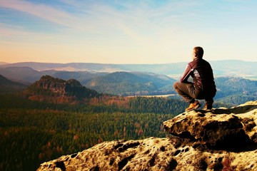 Young man in black sportswear is sitting on cliff edge and looking to misty valley bellow