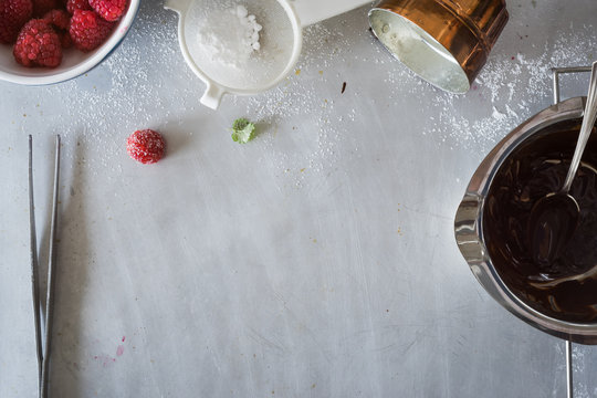Metal countertop surface with different staff for dessert plating: fresh raspberries, chocolate sauce, sugar powder, lemon balm