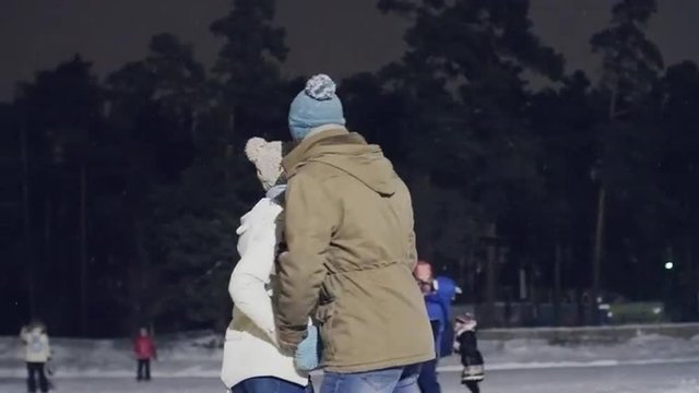 Happy young couple enjoying ice-skating and kissing at outdoor skating rink at night 