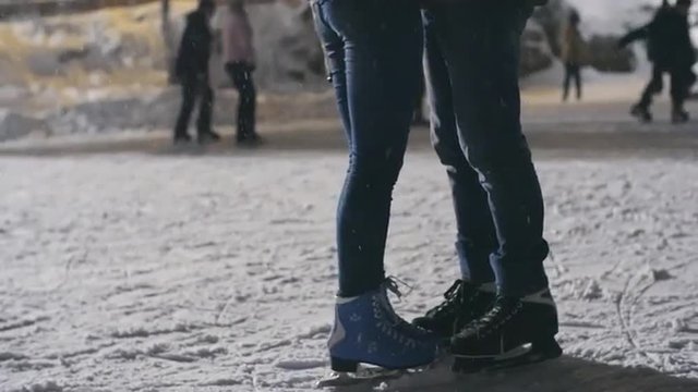 Low section of couple in ice skates hugging at outdoor skating rink, girl standing on tiptoes to kiss her boyfriend 