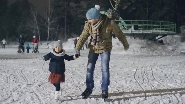 Positive young man teaching his lovely little daughter to ice skate at outdoor skating rink 