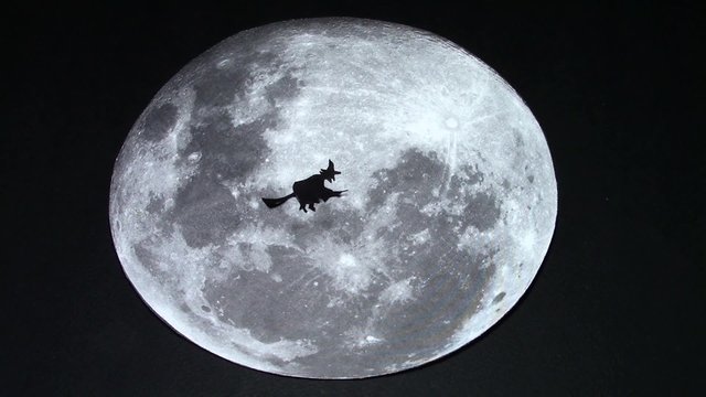 Paper cut witch silhouette slowly flying on her broom in front of the moon, going on her nightly workings