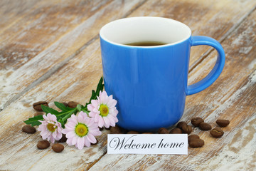 Welcome home card with mug of coffee and pink daisies on rustic wooden surface
