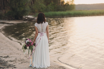 Fototapeta na wymiar Bride standing back to nature with a bouquet in the light of sunset