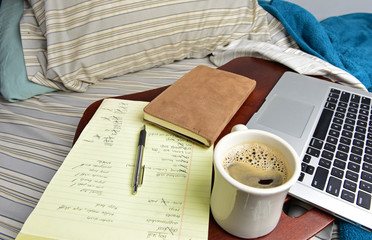 New Style Home Office/coffee mug, legal pad, pen, suede-bound journal, notebook computer on wood tray on unmade bed with wrinkled sheets and pillows in soft light