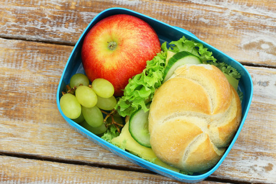 Lunch box containing bread roll with cheese, lettuce and cucumber, red apple on rustic wood
