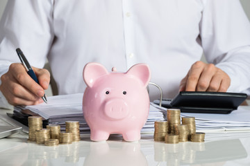 Businessman Calculating Invoice With Piggybank And Coins At Desk