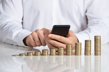 Businessman Using Smartphone With Stacked Coins At Desk