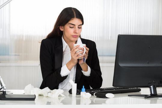 Sick Businesswoman Holding Cap Of Tee At Desk In Office