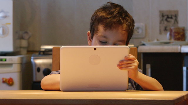 A cute little child holds at hands a white tablet PC at a table at home. Casual clothes
