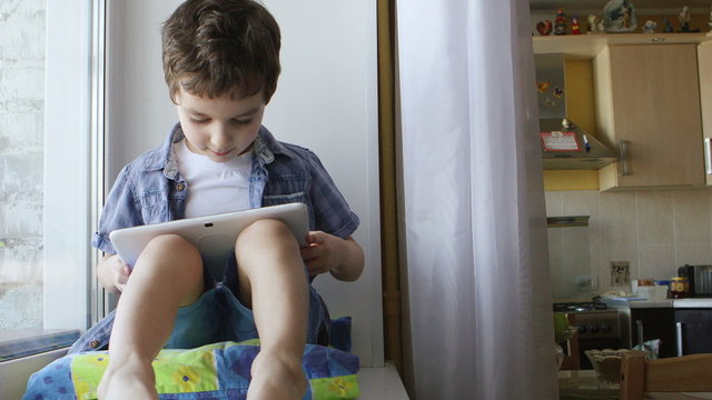 ZOOM: A cute little boy sits on a windowsill at home and touches a tablet PC
