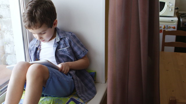 TOP VIEW: A cute little boy uses a white tablet PC on a windowsill at home
