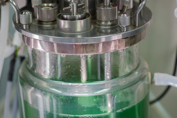 Close up of large glass beaker with green fluid, tubing and metallic parts for lab