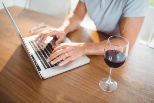 Focused senior man using laptop and drinking wine