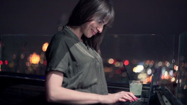 Woman using laptop and drinking cocktail on terrace at night

