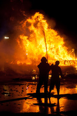 Fototapeta premium Silhouette of Firemen fighting a raging fire with huge flames of burning timber