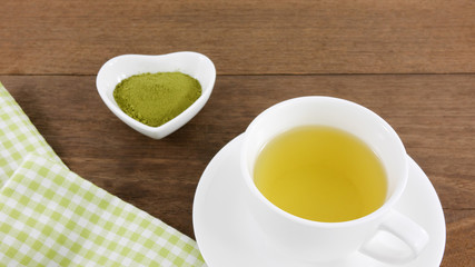 The Japanese matcha green tea powder on ceramic heart shaped bowl and cup of hot green tea with cotton fabric on wooden planks.