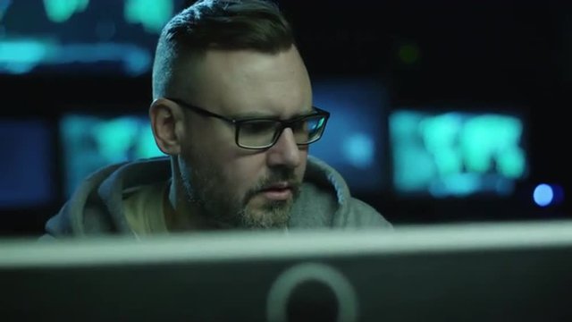 Portrait footage of concentrated male employee working on a computer in a dark office room with display screens with maps and data. Shot on RED Cinema Camera.