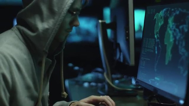 Two hackers in hoods work on a computers with maps and data on display screens in a dark office room. Shot on RED Cinema Camera.