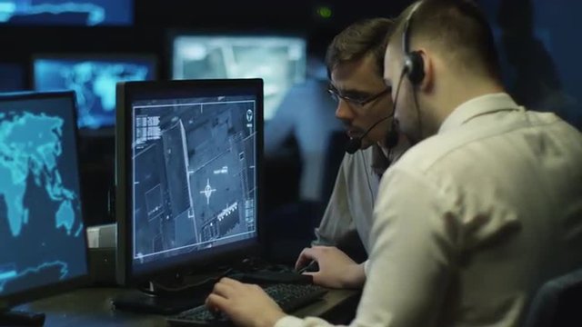 Two IT professionals are spying on businessman through satellite surveillance on computer in a dark monitoring room filled with display screens. Shot on RED Cinema Camera.