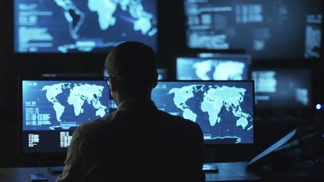 Male IT employee comes to monitoring room to work on computer with maps and data on display in a dark office room. Shot on RED Cinema Camera.