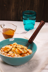 Oatmeal porridge with banana slices, cinnamon and caramel sauce over white wooden table. Selective focus