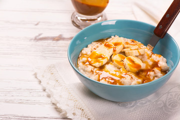 Oatmeal porridge with banana slices, cinnamon and caramel sauce over white wooden table. Selective focus