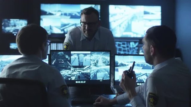 Group of security officers are having a conversation at work in a dark monitoring room filled with display screens. Shot on RED Cinema Camera.