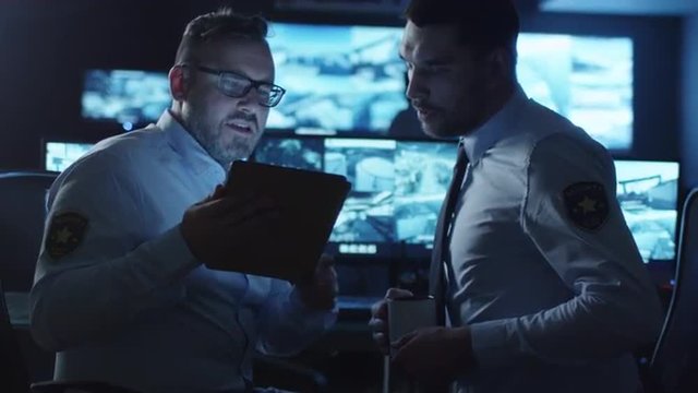 Security officer is using a tablet computer in a dark monitoring room filled with display screens. Shot on RED Cinema Camera.