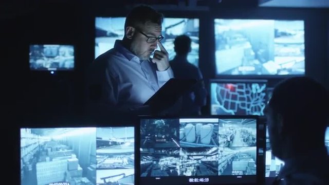 Security officer is using a tablet computer in a dark monitoring room filled with display screens. Shot on RED Cinema Camera.