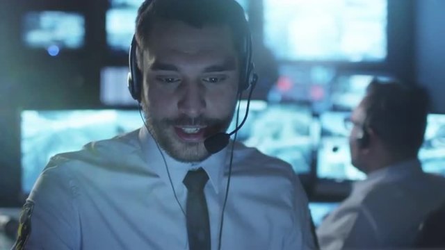 Technical support specialist is talking on a headset while working on a computer in a dark monitoring room filled with display screens. Shot on RED Cinema Camera.