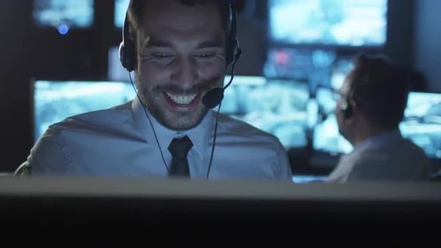 Technical support specialist is talking on a headset while working on a computer in a dark monitoring room filled with display screens. Shot on RED Cinema Camera.