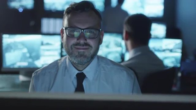 Happy office employee in white shirt is sitting next to a computer, looking at the camera and smiling in a dark monitoring room filled with display screens. Shot on RED Cinema Camera.