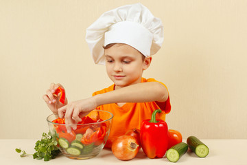 Little boy in chefs hat puts chopped vegetables for a salad in a bowl