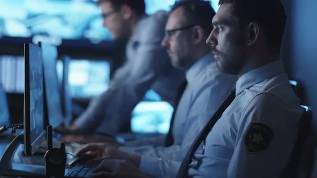 Group of security officers are working in a dark monitoring room filled with display screens. Shot on RED Cinema Camera.