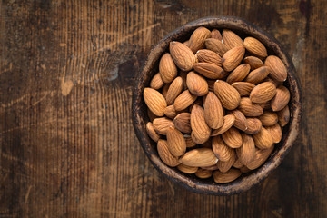 Almonds in brown bowl on textured wooden background, top view. Copy space on left side