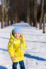 Cute little girl wearing yellow jacket and blue hat playing with snowball in winter park on sunny day