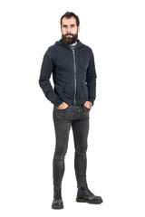 Tough confident stylish punker in black hooded sweatshirt looking at camera with hands in pockets. Full body length portrait isolated over white studio background.
