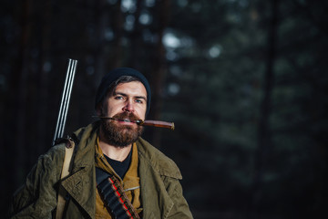 brutal hunter, bearded man in warm hat with a gun in his hand, a knife a backpack and smoking pipe in the dark wild forest in the autumn