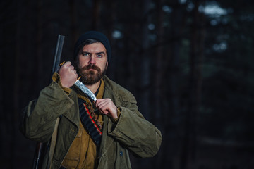 brutal hunter, bearded man in warm hat with a gun in his hand, a knife a backpack and smoking pipe in the dark wild forest in the autumn