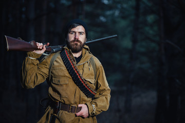Fototapeta premium brutal hunter, bearded man in warm hat with a gun in his hand, a knife a backpack and smoking pipe in the dark wild forest in the autumn