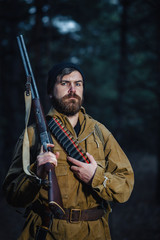 brutal hunter, bearded man in warm hat with a gun in his hand, a knife a backpack and smoking pipe in the dark wild forest in the autumn