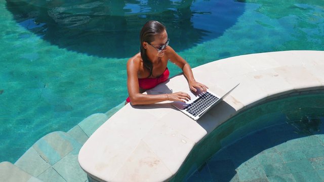Woman working on her laptop computer sitting at poolside. Beautiful young female model working freelance at swimming pool on laptop.