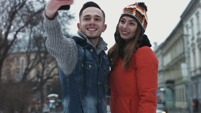 Sweet young couple making selfie on smartphone. Male and female in their 20s having a date outdoors and taking pictures by using mobile device. People make faces on camera.  