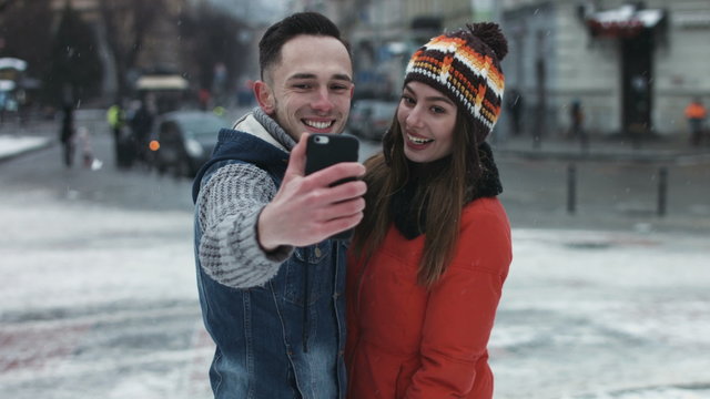 Young attractive couple making faces on camera. Man and woman taking photographs by using smartphone. Boy and girl make selfie outdoors in the middle of the european old town while its snowing. 